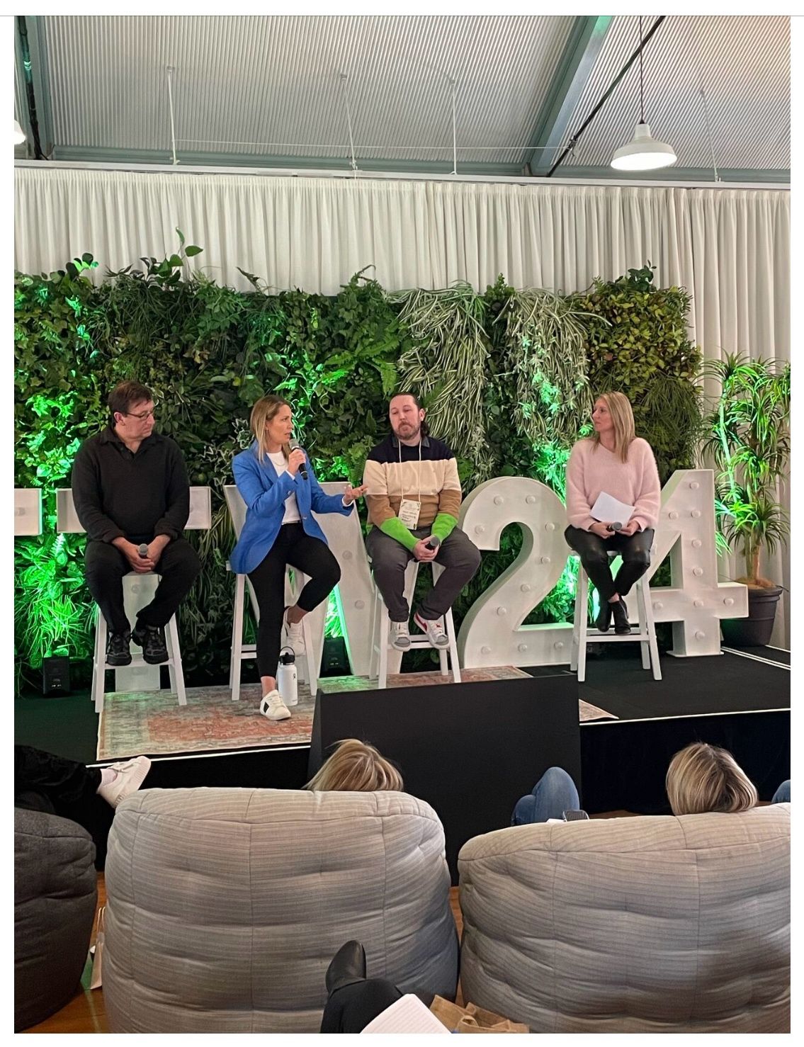 Natalie and three panelists sitting on high stools against a backdrop of over sized numbers 2024. Attendees are sitting in lounge chairs.